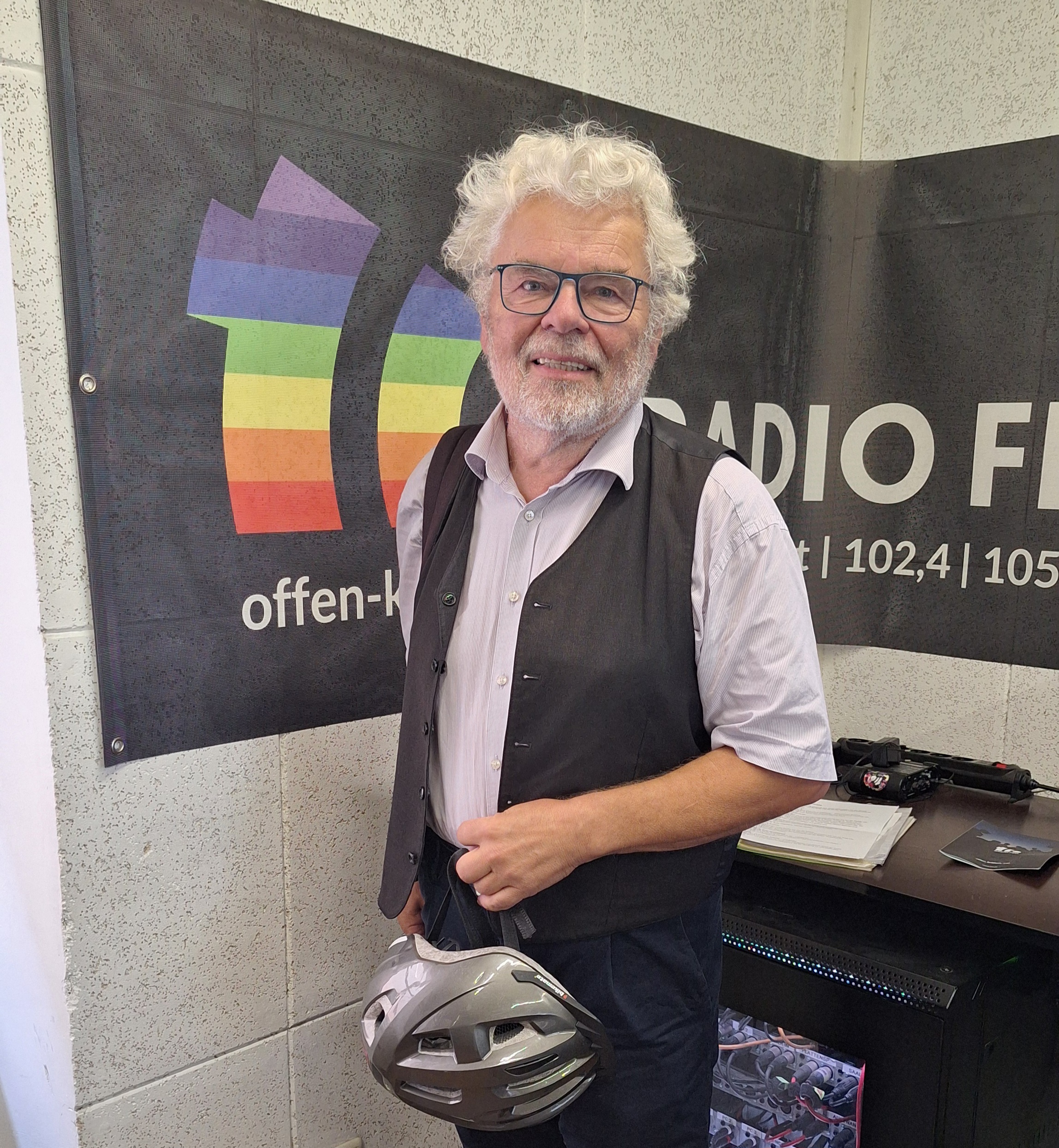 Ein Mann steht im Radio FRO Studio und lächelt in die Kamera. Er hält einen Fahrradhelm in der Hand und hinter ihm hängt ein regenbogenfarbenes Radio FRO Banner an der Wand.