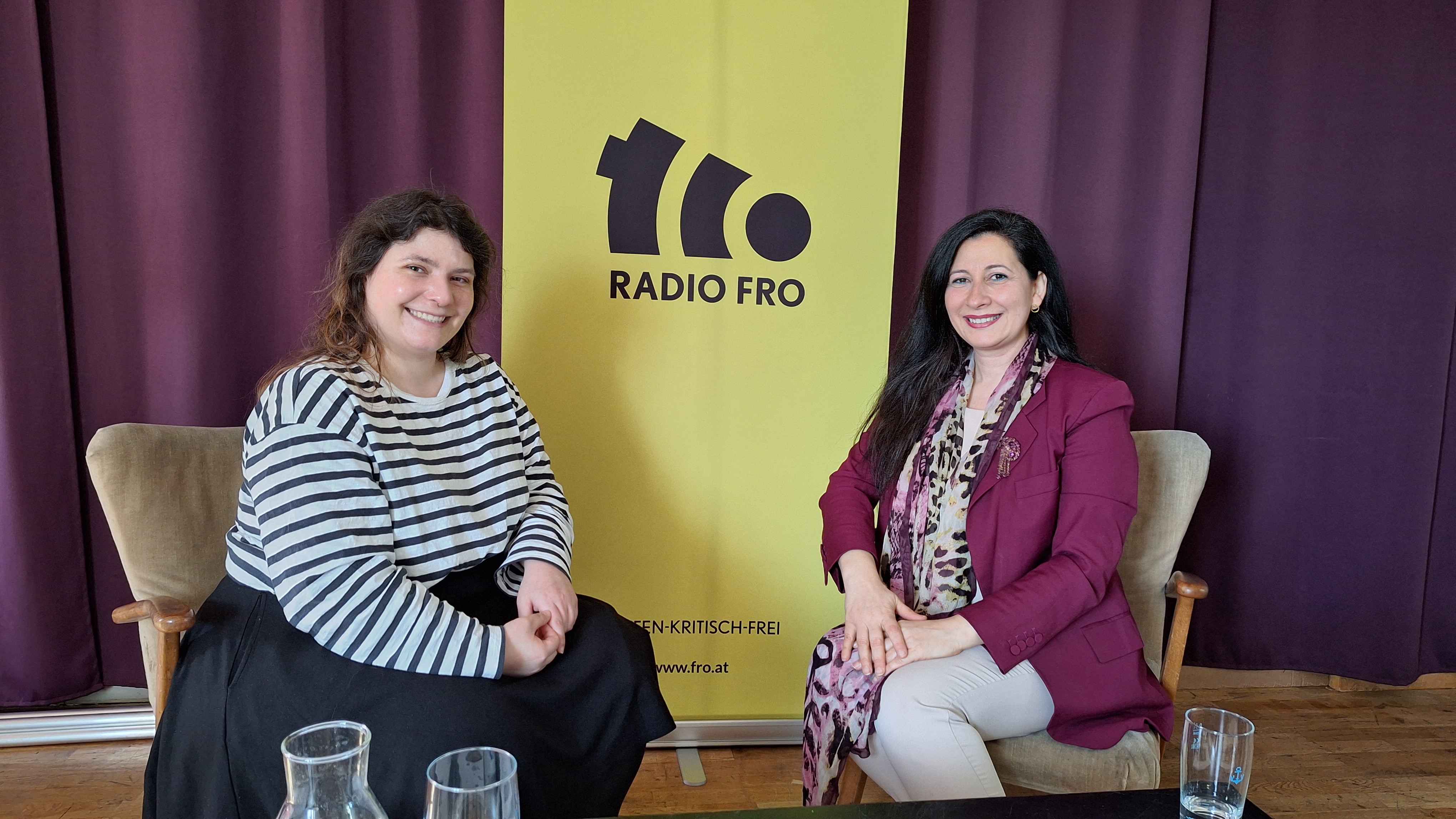 2 Frauen sitzen auf Sesseln. Hinter ihnen ist ein violetter Vorhang und ein gelbes Roll-up, auf dem das Radio FRO Logo in schwarz zu sehen ist. Beide lächeln in die Kamera