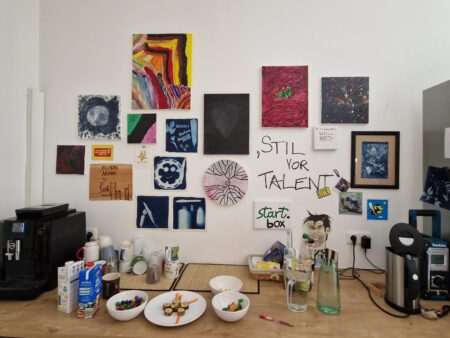 Häferl und Teller stehen auf einem Tisch herum. An der Wand dahinter sind selbstgemalte Bilder zu sehen. 