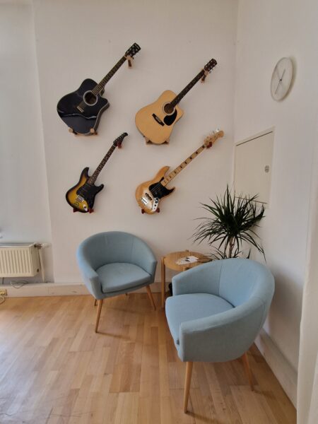 Es sind zwei stoffsessel zu sehen. Sie stehen auf einem Holzboden und hinter ihnen an der Wand hängen Gitarren.